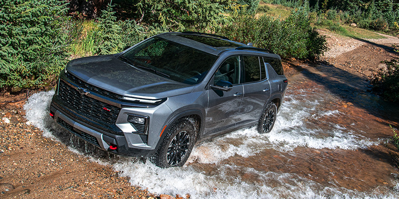 A gray Chevrolet Traverse crossing a stream with water splashing around its tires.