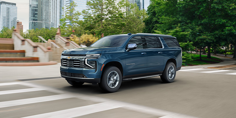 Front-three-quarter view of a dark blue Chevrolet Suburban driving on a city street past green trees and modern skyscrapers.