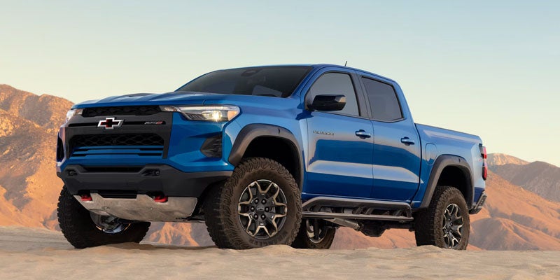 A bright blue Chevrolet Colorado ZR2 pickup truck parked on desert sand with mountains in the background.