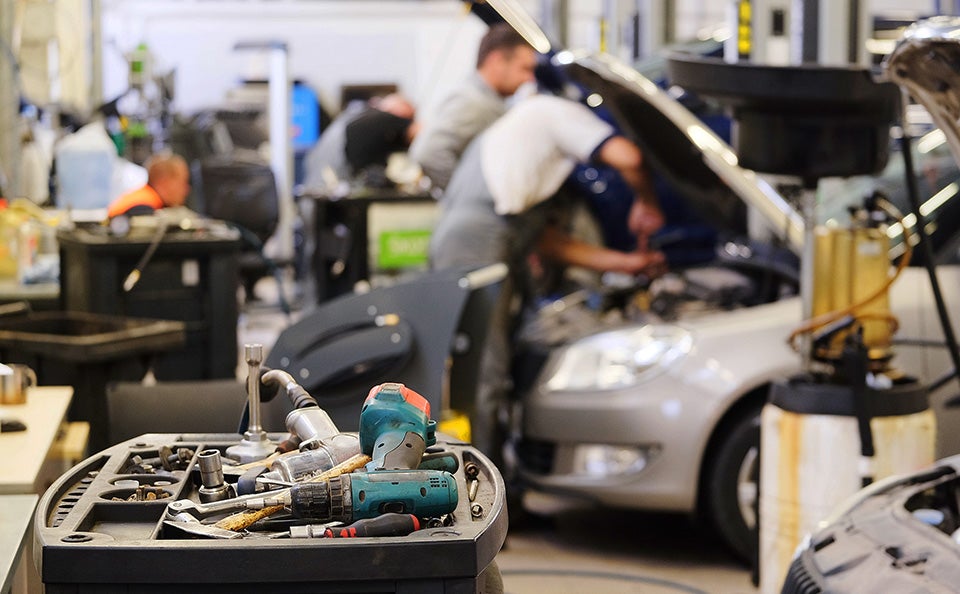 A detailed tool cart in the foreground with mechanics working under the open hood of a car in a busy repair shop background.