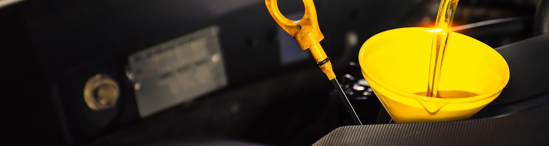 A stream of clean golden engine oil being poured through a bright yellow funnel into a car engine, with a yellow oil dipstick handle visible nearby.