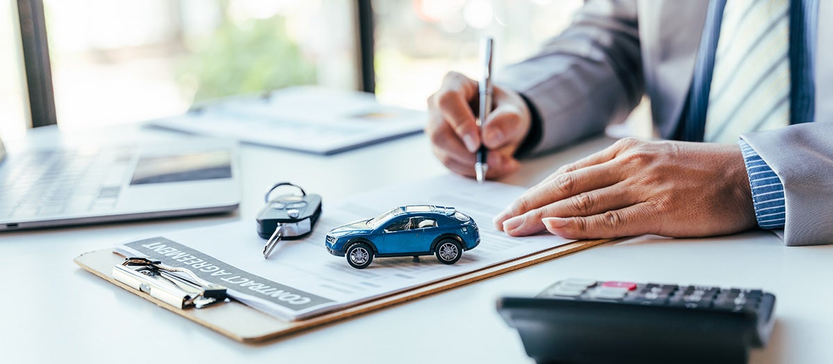 Close-up of a person signing a car contract agreement next to a blue miniature toy car, key fob, and calculator.